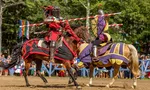 Two knights in colored armor jousting on horseback during a medieval festival with spectators behind a wooden fence.