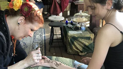 Artist creating an intricate henna tattoo on a young woman's forearm at a vibrant outdoor market.