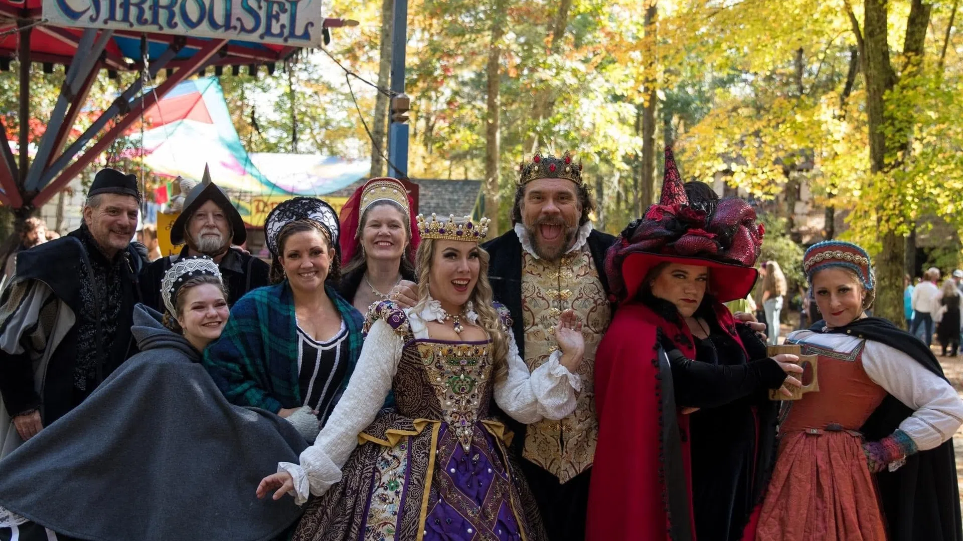 Group of people dressed in colorful medieval costumes posing happily in front of a carousel in autumn.