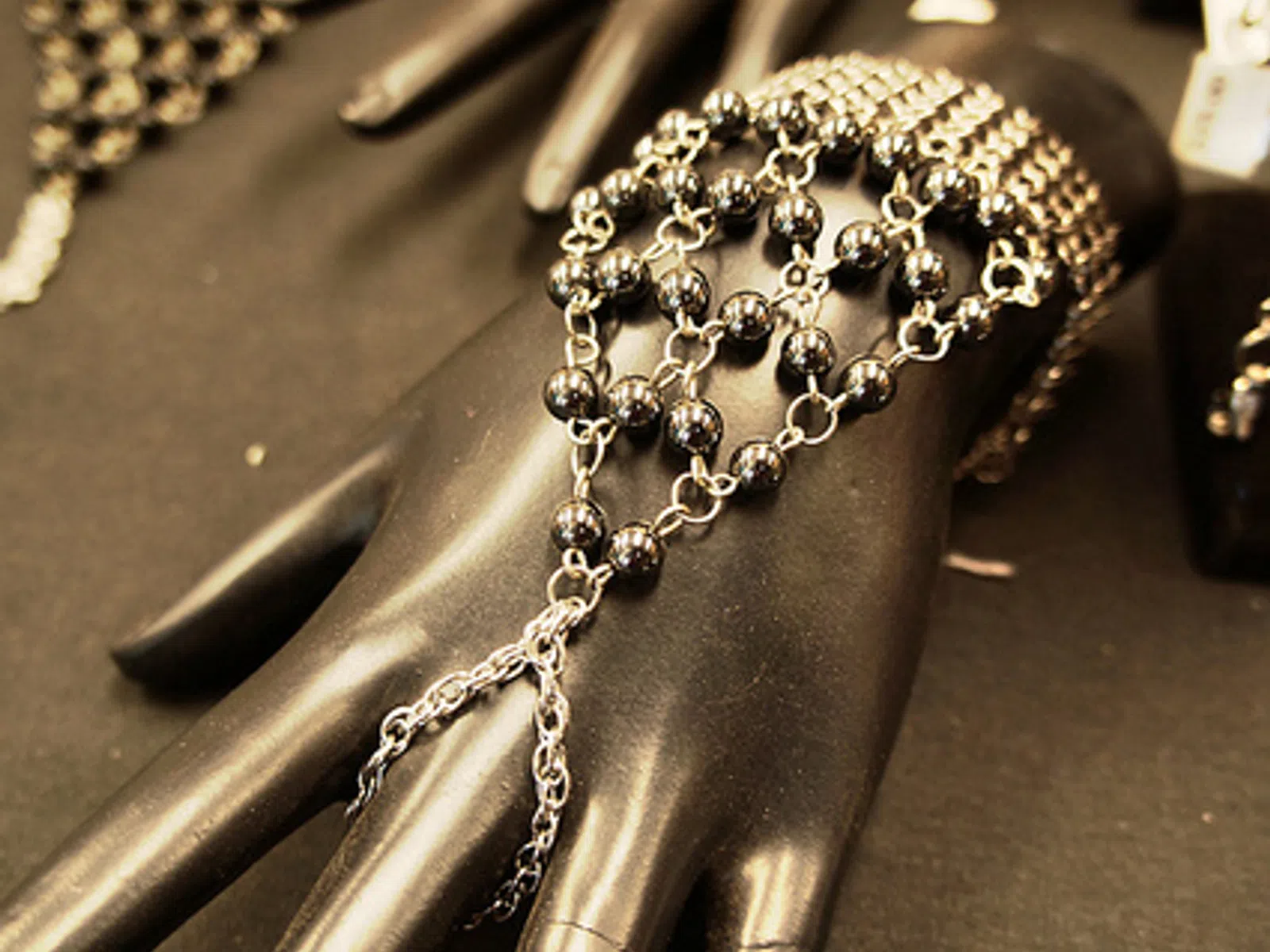 Black mannequin hand displaying a silver and black bead chain hand bracelet on a dark surface.