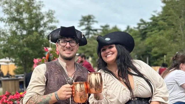 Smiling couple dressed in pirate costumes clinking copper mugs while sitting outdoors with flowers and trees.