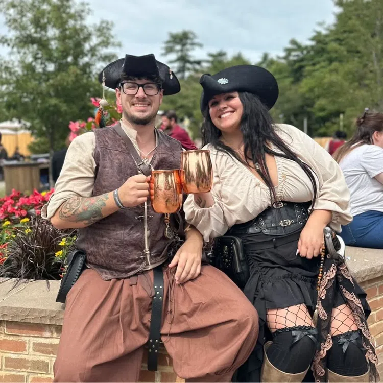 Smiling couple dressed in pirate costumes clinking copper mugs while sitting outdoors with flowers and trees.