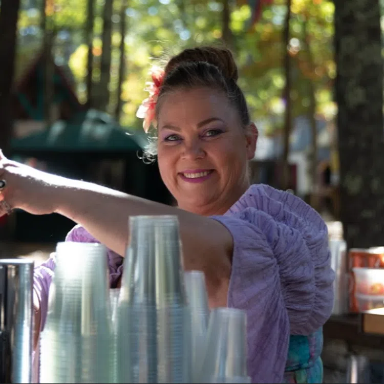 Smiling woman in purple blouse pouring a seasonal beer at an outdoor autumn festival with plastic cups and trees in background