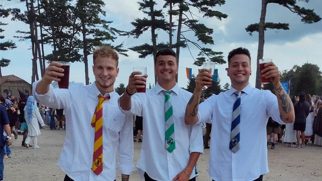 Three young men in white shirts and colorful ties raise drinks outdoors at a festive gathering with trees and crowd.