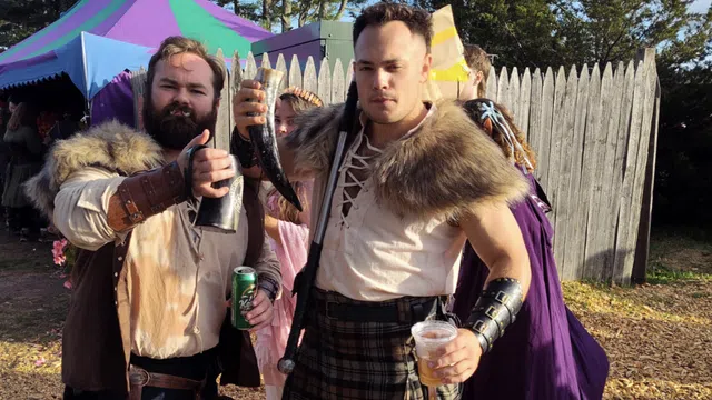 Two men in medieval costumes holding drinking horns and cups at a Renaissance fair with colorful tents and wooden fences.