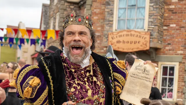 Man dressed as a medieval king in purple and gold robes holding a pewter mug and a parchment in a Renaissance fair setting