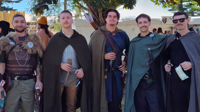 Group of five men dressed in medieval costumes with capes, swords, and props at an outdoor festival under trees.